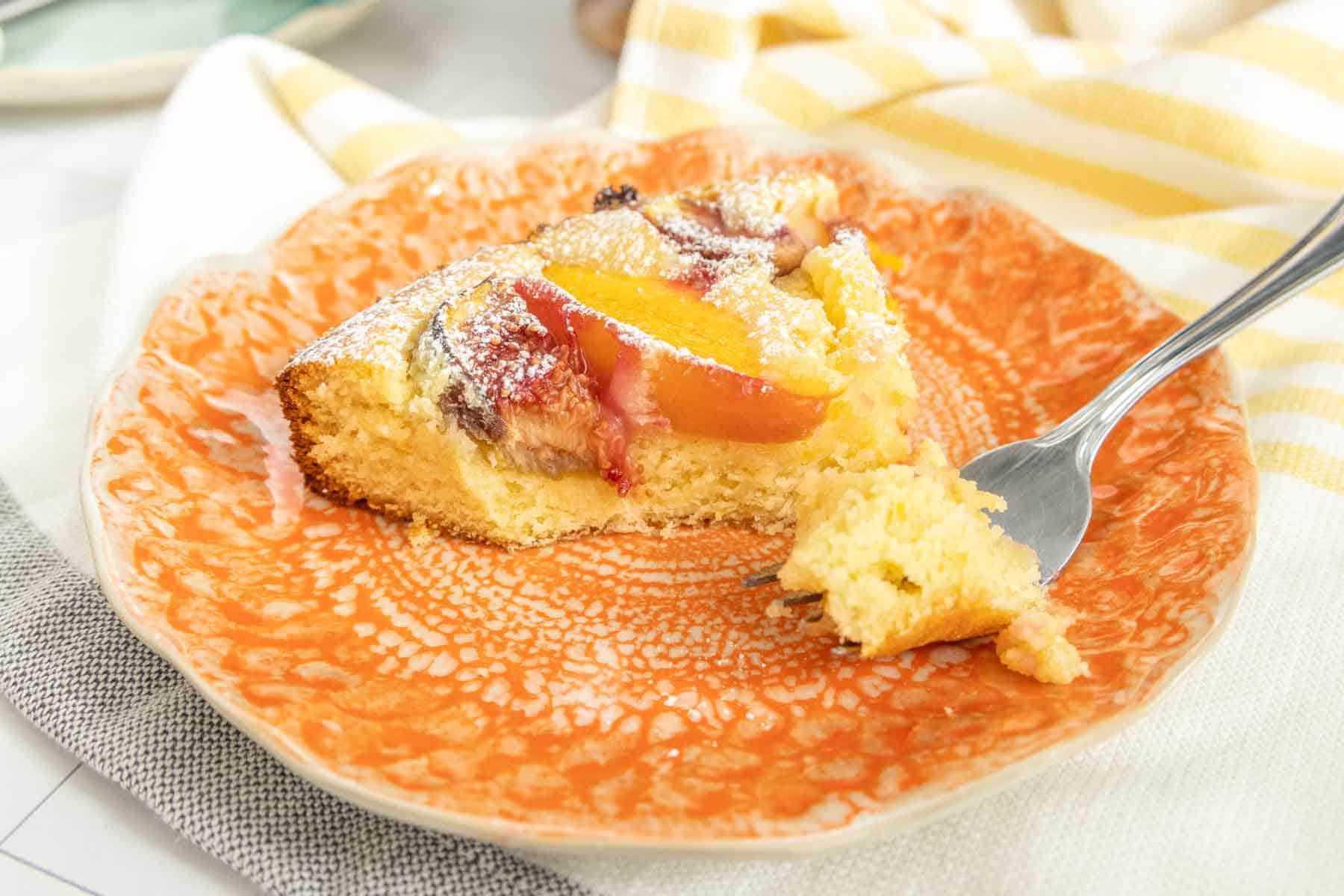 A close-up of a slice of figcake on an orange plate with a fork resting on the side. The cake appears to be soft and moist, topped with powdered sugar and nectarine slices.