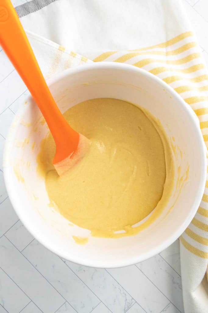 A white mixing bowl with yellow cake batter and an orange spatula inside, placed on a striped kitchen towel.