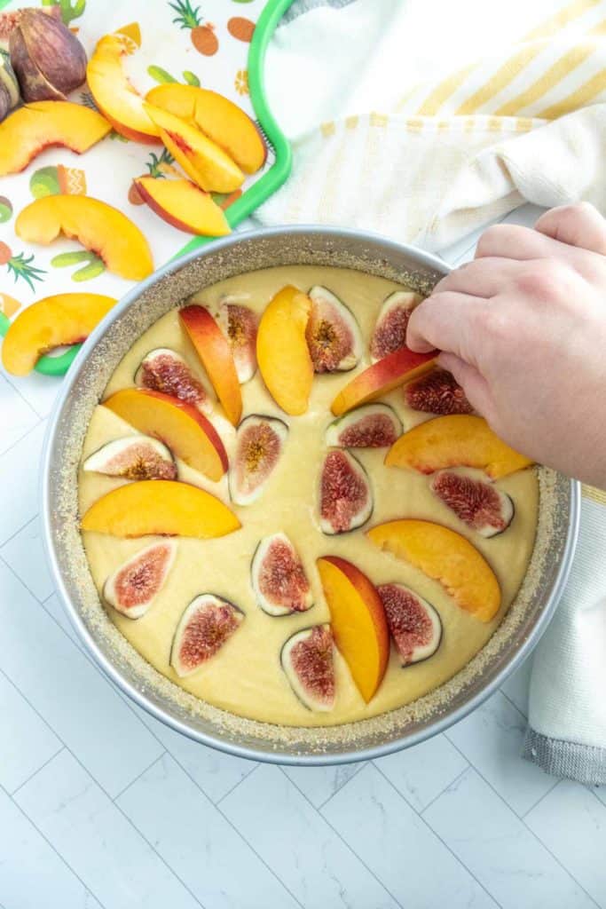 A round baking pan with fig and nectarine slices being placed on cake batter. A person's hand is arranging slices. Fruit slices and a towel are visible in the background.