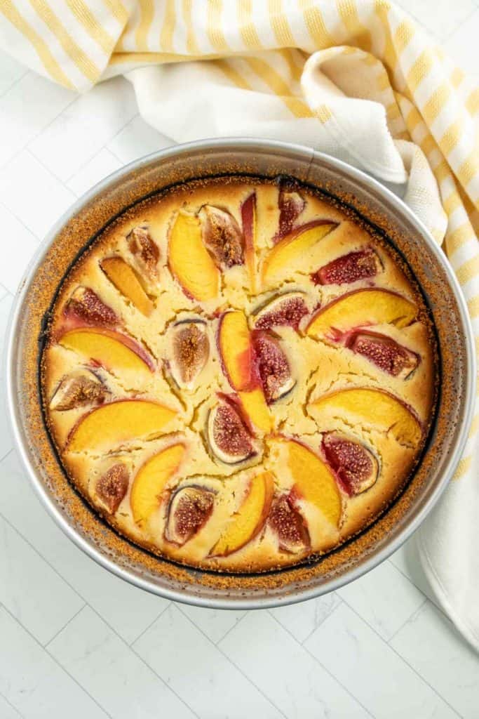 A round cake in a pan topped with sliced nectarines and figs, placed on a light-colored surface with a yellow and white striped cloth in the background.