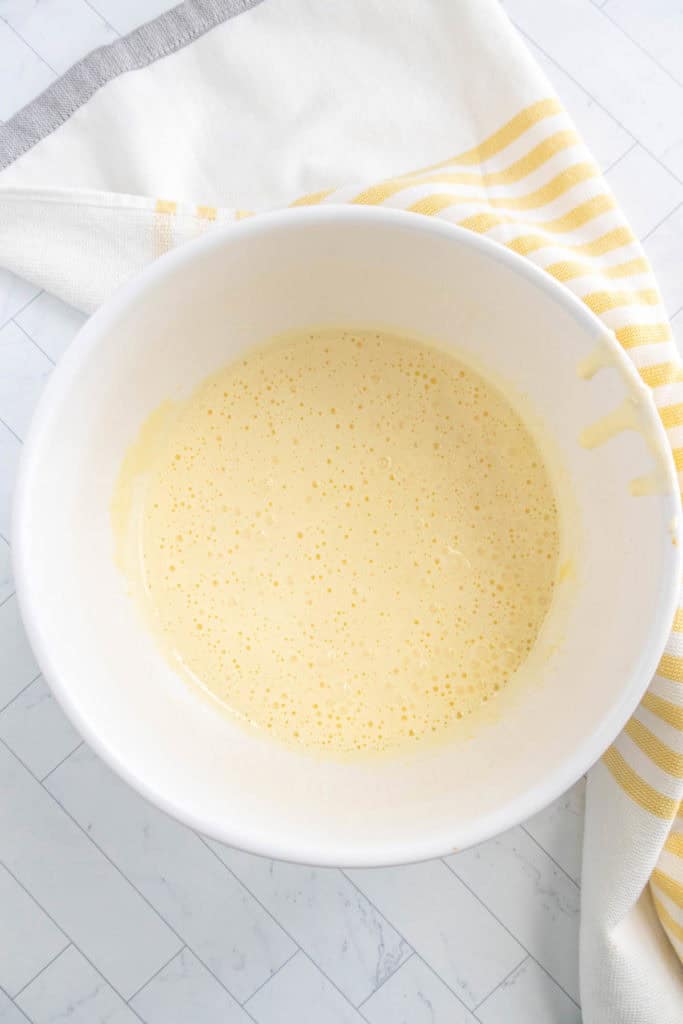 A white bowl filled with a light yellow batter is placed on a white and gray countertop next to a folded white and yellow striped cloth.