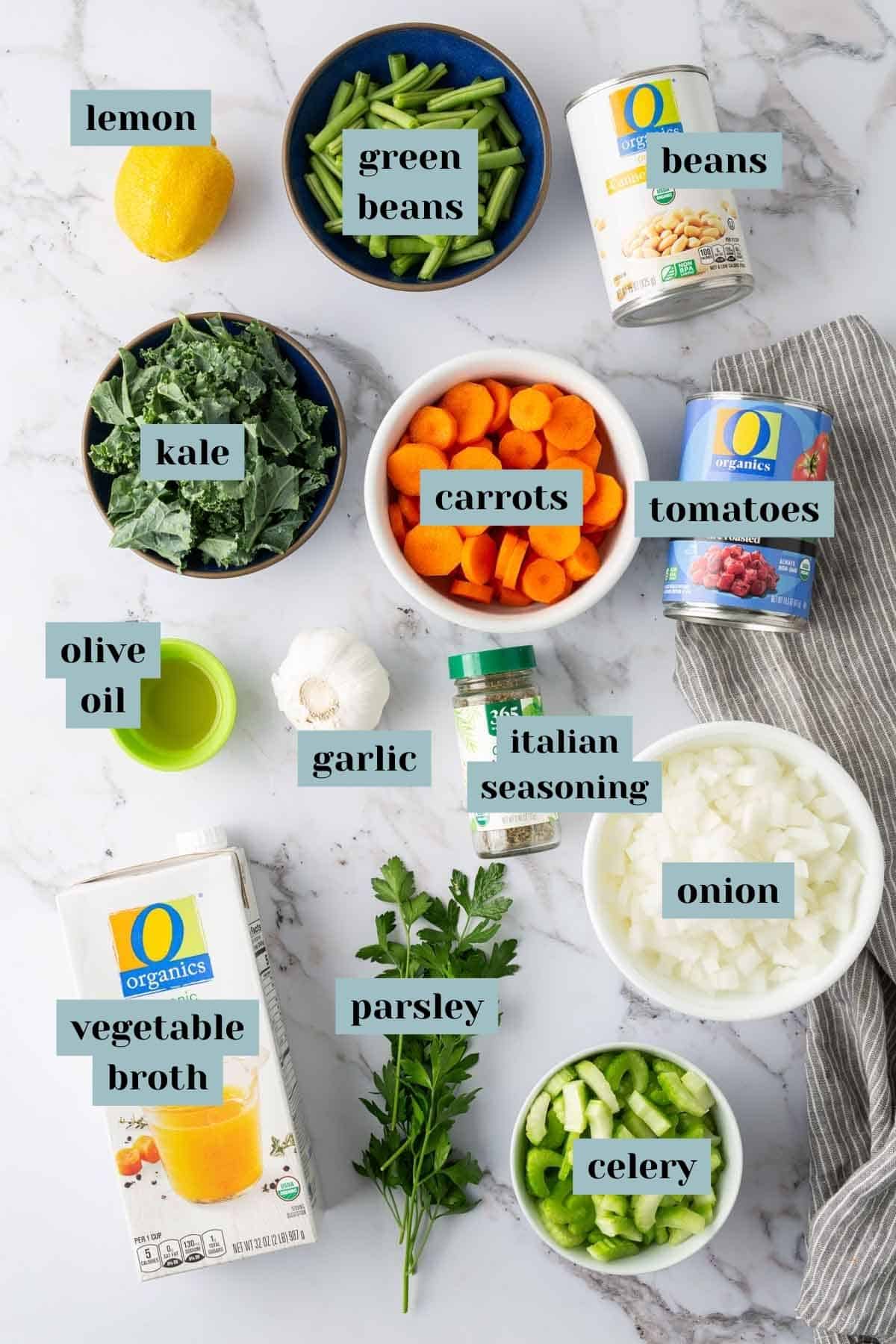 A flat lay image shows various labeled ingredients on a white marble surface, including lemon, green beans, canned beans, kale, carrots, canned tomatoes, olive oil, garlic, Italian seasoning, onion, vegetable broth, parsley, and celery.