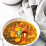 A bowl of vegetable soup containing carrots, beans, tomatoes, and herbs sits on a white surface with another bowl in the background, next to a grey-striped cloth.