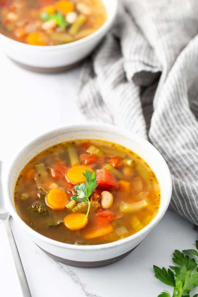 A bowl of vegetable soup containing carrots, beans, tomatoes, and herbs sits on a white surface with another bowl in the background, next to a grey-striped cloth.