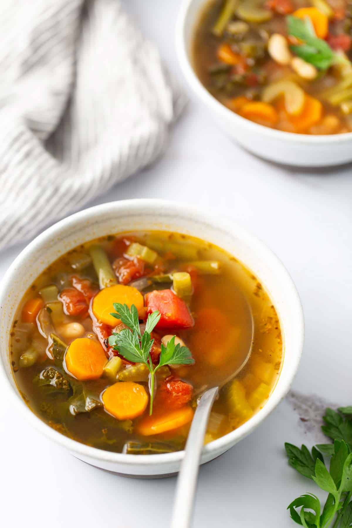 Two bowls of vegetable soup with carrots, green beans, and tomatoes garnished with parsley, with a striped napkin in the background.