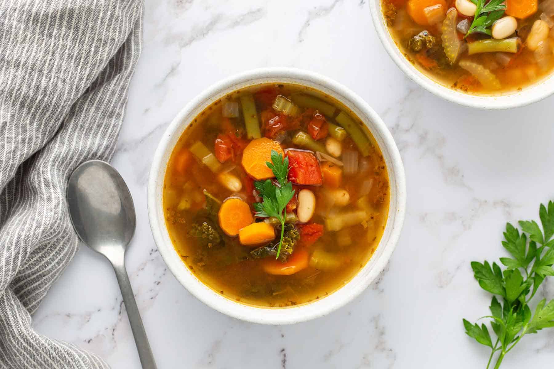 A bowl of vegetable soup with carrots, tomatoes, beans, and parsley garnish, placed on a marble surface beside a gray-striped cloth and a spoon. A sprig of parsley lies next to the bowl.