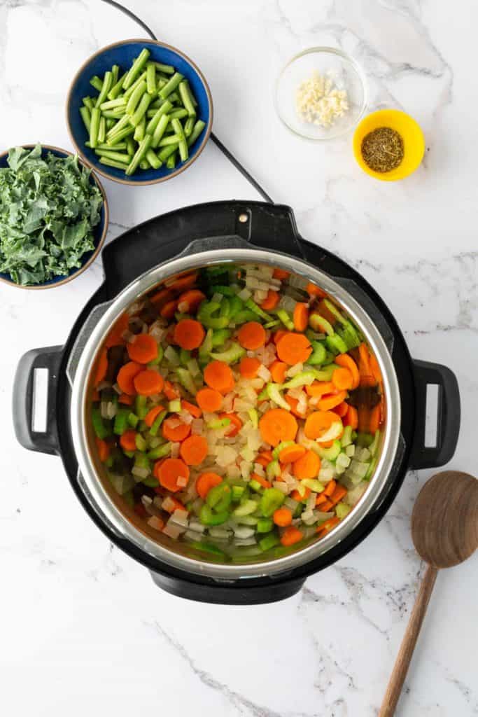 An overhead view of a pot with chopped carrots, celery, and onions. Surrounding the pot are small bowls containing chopped kale, green beans, minced garlic, and a spice mix, and a wooden spoon.