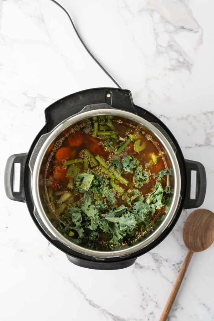 A top view of a pressure cooker filled with a vegetable soup containing greens, carrots, and other vegetables on a marble surface. A wooden ladle lies to the right of the cooker.