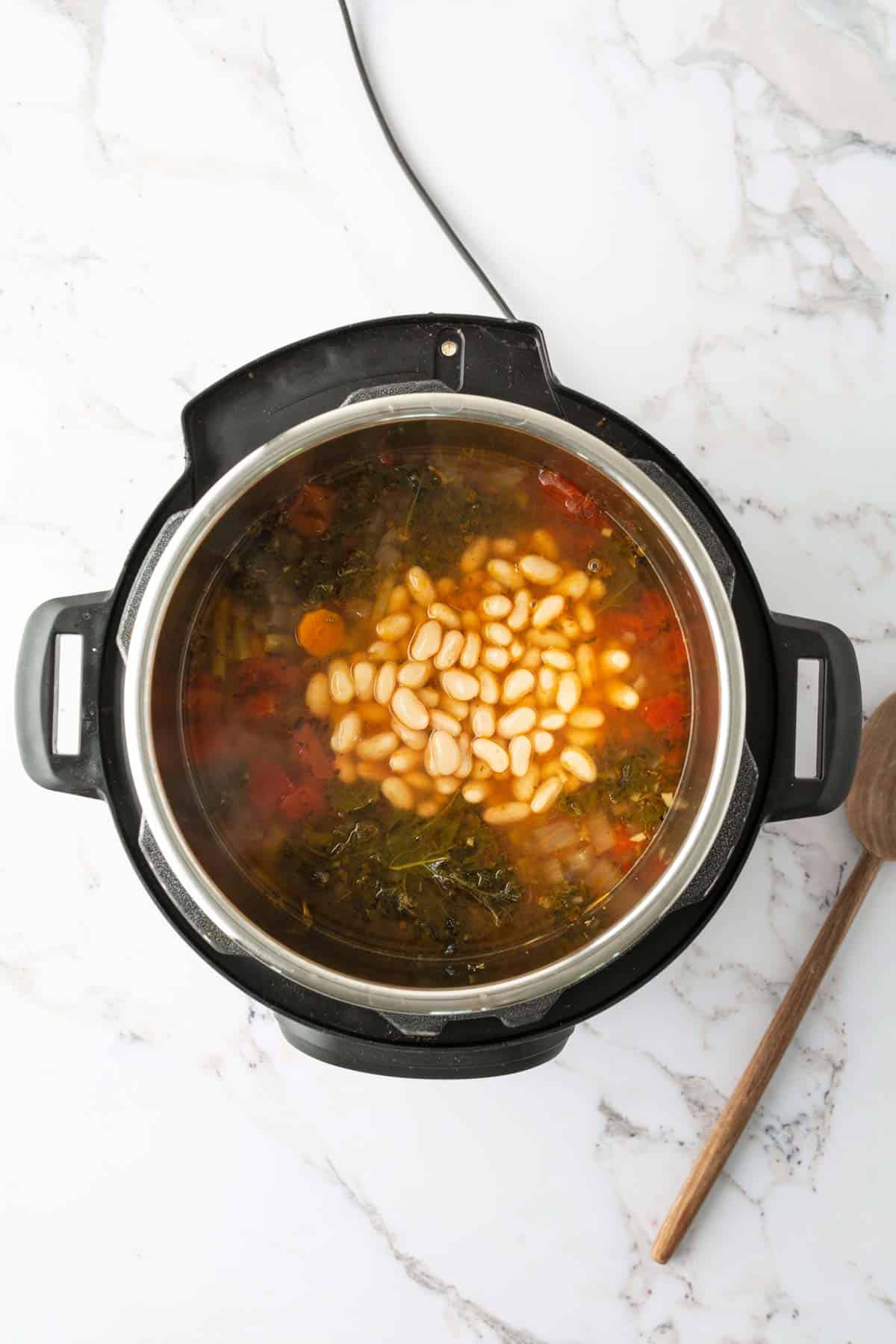 An overhead view of a multi-cooker filled with a broth containing white beans, vegetables, and greens, on a marble countertop next to a wooden spoon.