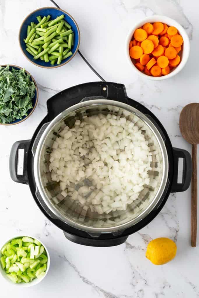 An overhead view of a pressure cooker filled with chopped onions, surrounded by bowls of sliced green beans, carrots, greens, celery, and a lemon on a marble countertop.