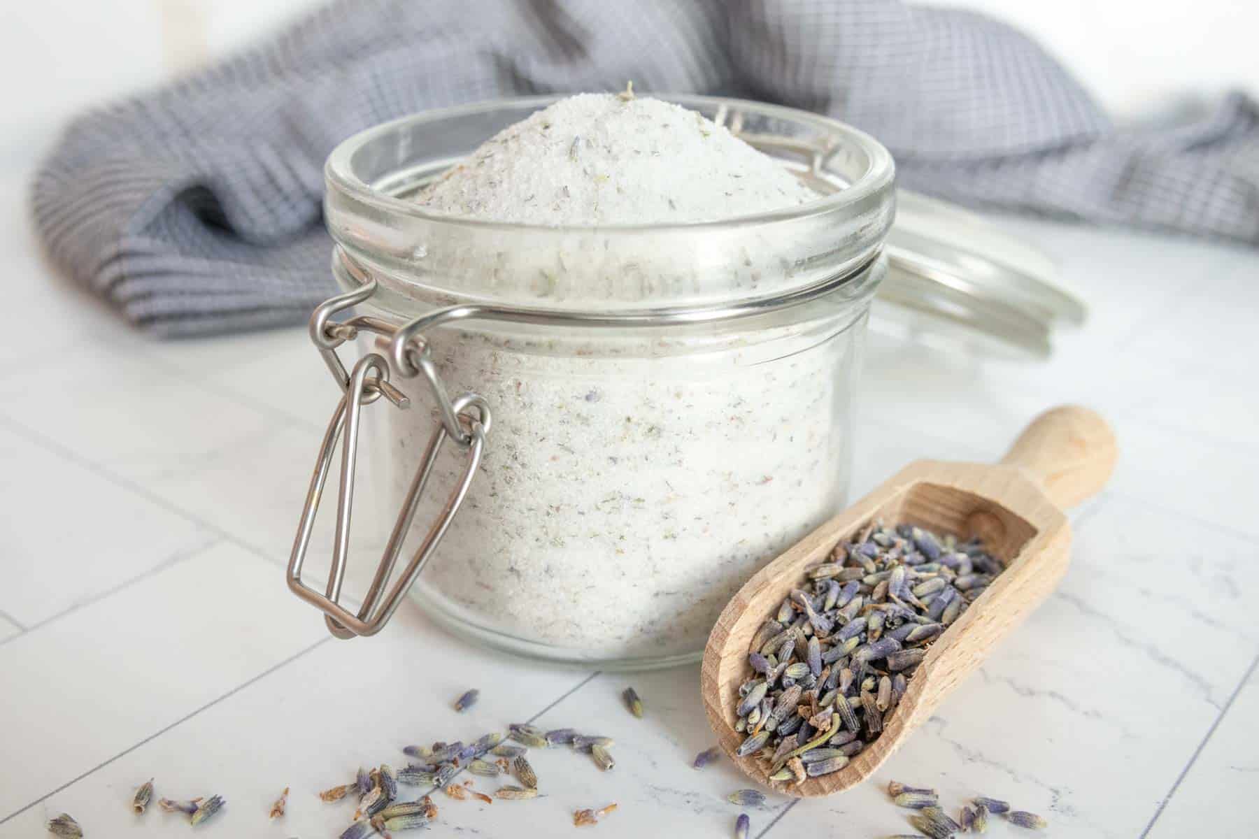 A glass jar filled with lavender sugar and a small wooden scoop with dried lavender buds positioned on a white tiled surface. A checkered cloth is partially visible in the background.