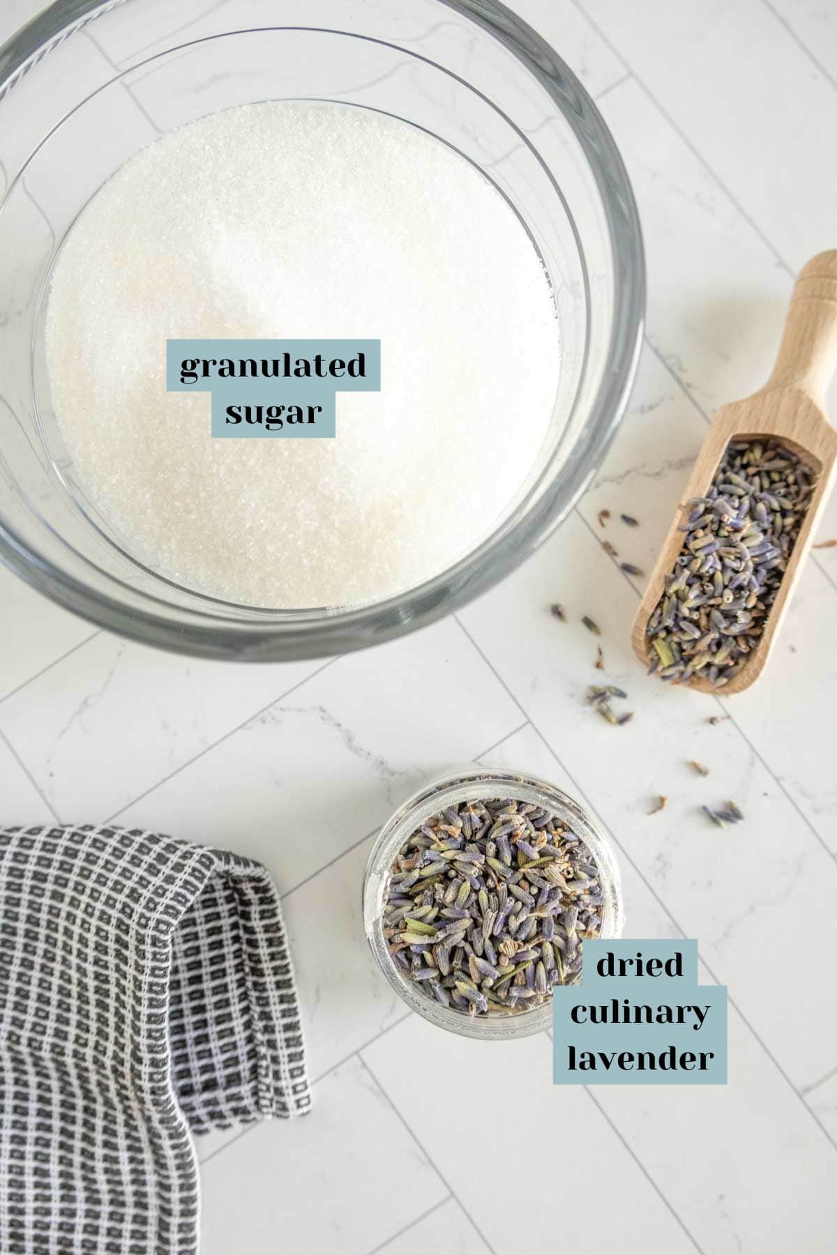 A glass bowl of granulated sugar and a small scoop of dried culinary lavender next to a jar filled with lavender buds on a light-colored surface with a cloth nearby.