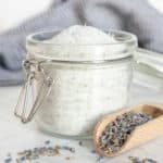 A glass jar filled with sugar beside a wooden scoop and scattered lavender buds, with a cloth in the background.