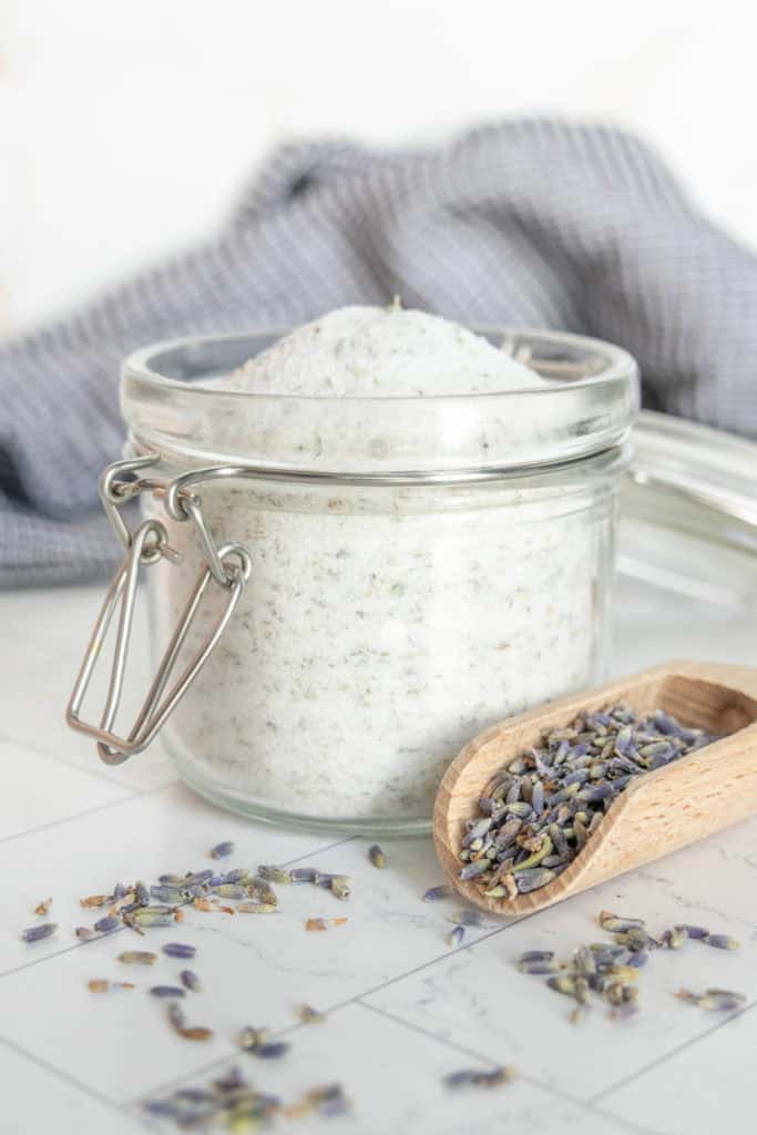 A glass jar filled with sugar beside a wooden scoop and scattered lavender buds, with a cloth in the background.