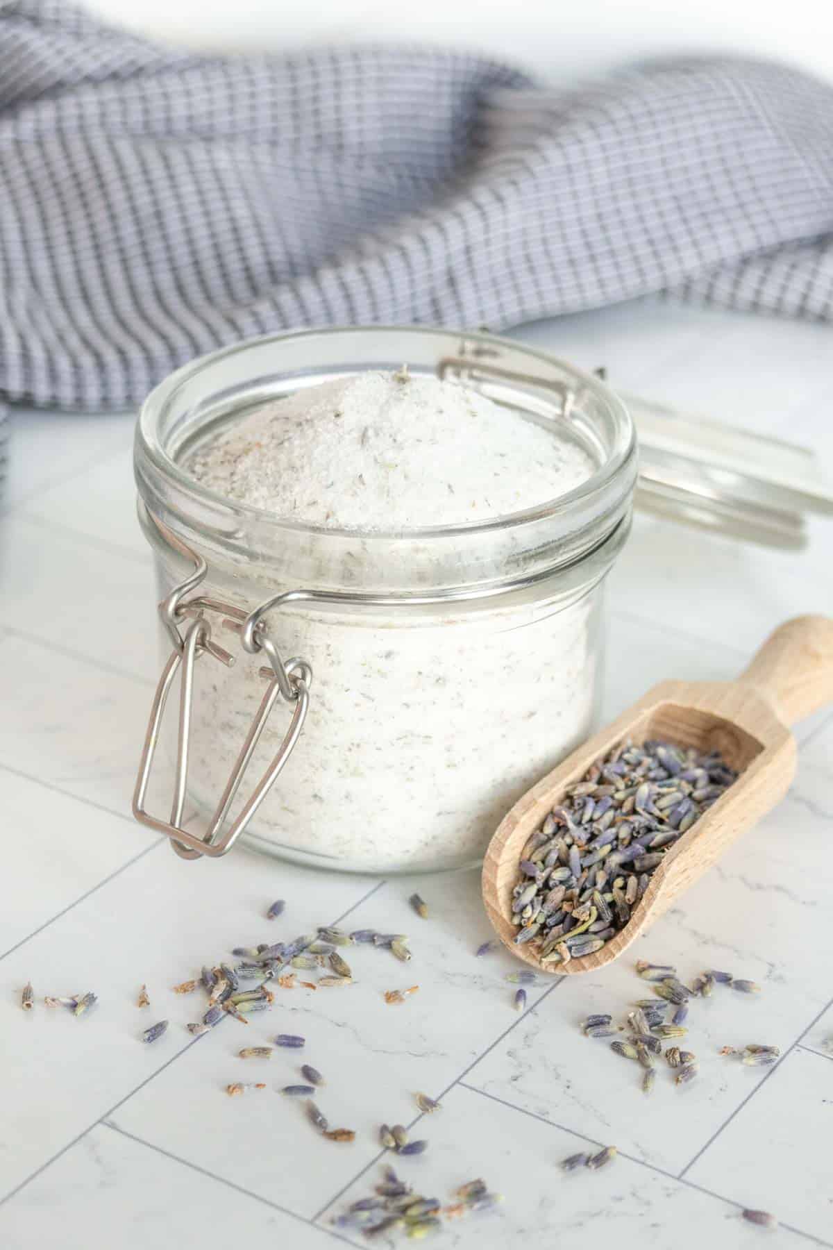 A jar of sugar sits on a tiled surface next to a wooden scoop filled with dried lavender buds. A gray cloth is folded in the background.