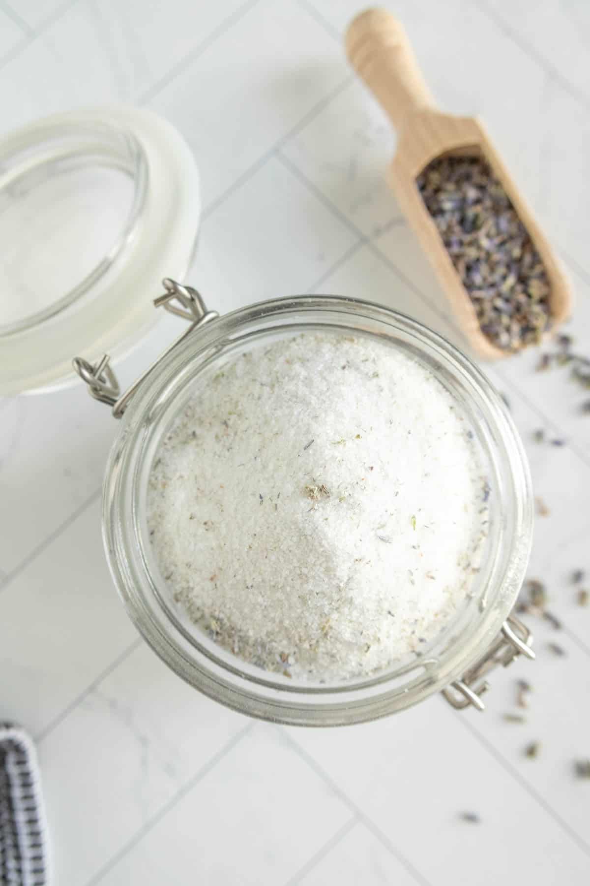 A glass jar filled with lavender sugar next to a wooden scoop containing dried lavender, all placed on a light-colored tiled surface.