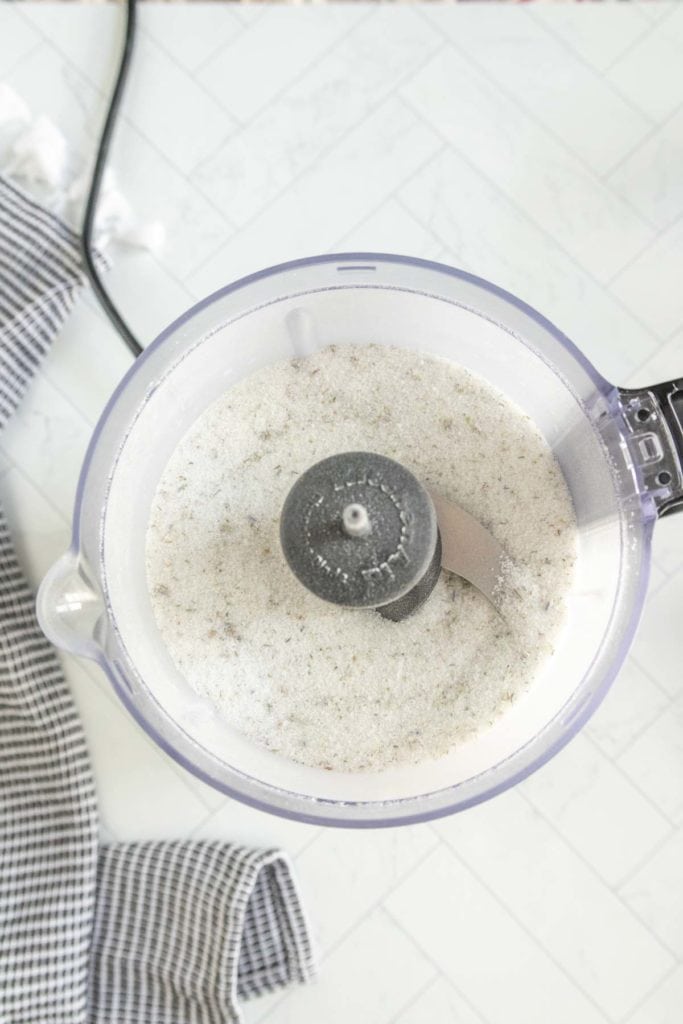 A food processor viewed from above, containing a white powdered mixture. A portion of a black and white checkered cloth is visible on the counter next to it.