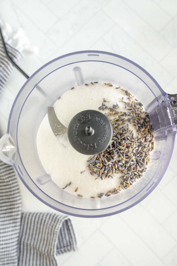 Top view of a food processor bowl containing sugar and dried lavender buds, with part of a striped cloth visible beside it.