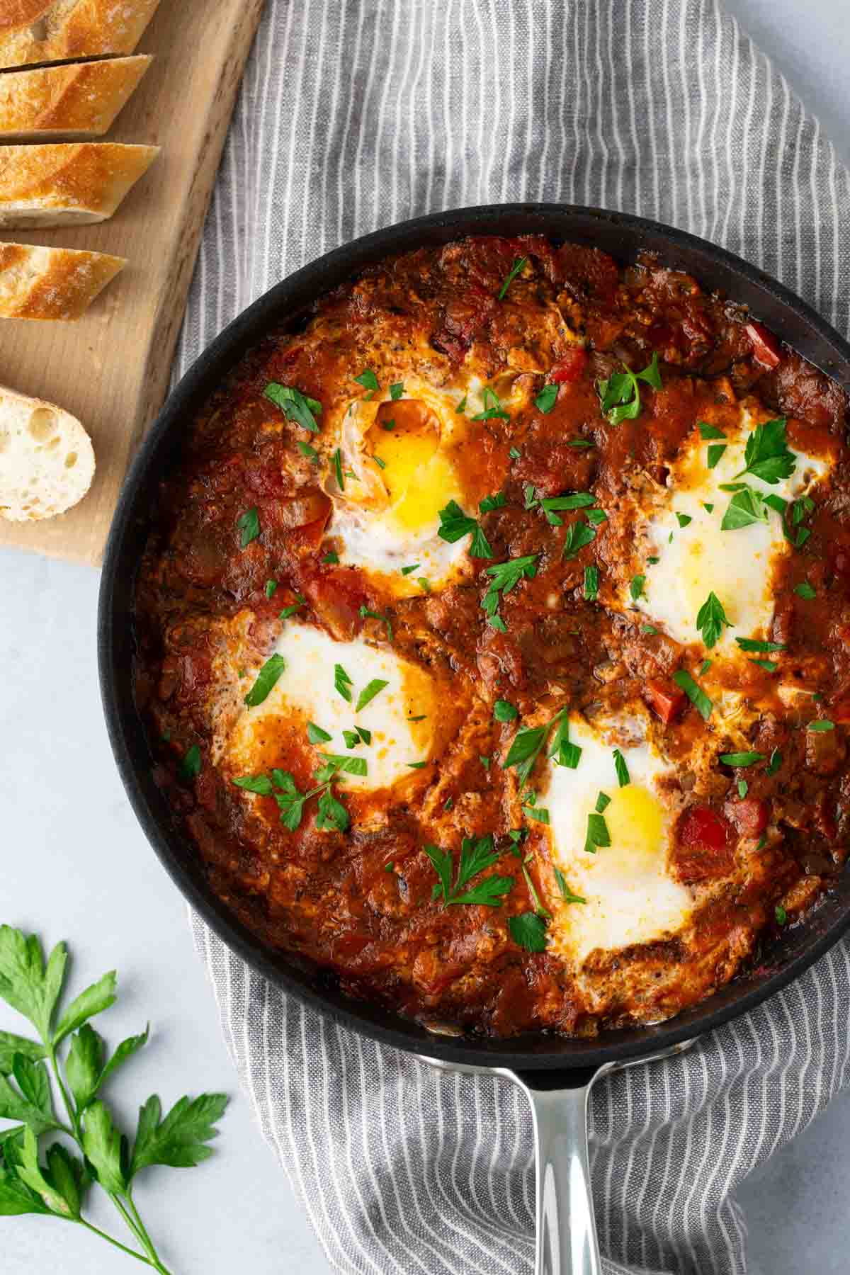 A black skillet with shakshuka, featuring eggs poached in a tomato and pepper sauce, garnished with chopped parsley. Slices of baguette are on a cutting board nearby.