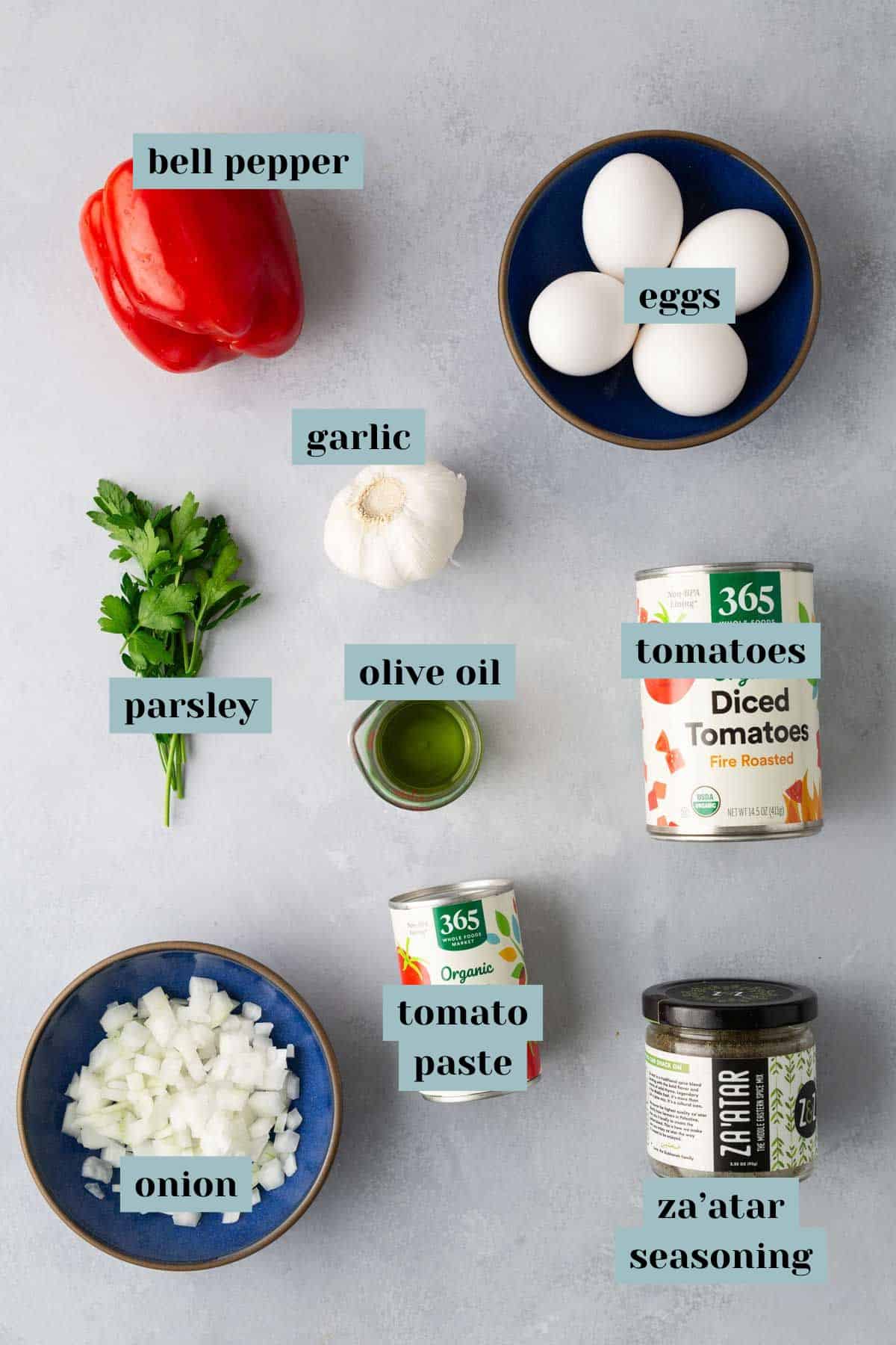 A flat lay of cooking ingredients labeled with tags. Items include a bell pepper, garlic, parsley, onion, eggs, tomato paste, a can of diced tomatoes, olive oil, and za'atar seasoning.