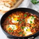 A skillet with shakshuka, a dish of poached eggs in a tomato and pepper sauce, garnished with parsley. Slices of bread and a bowl of sauce are in the background.