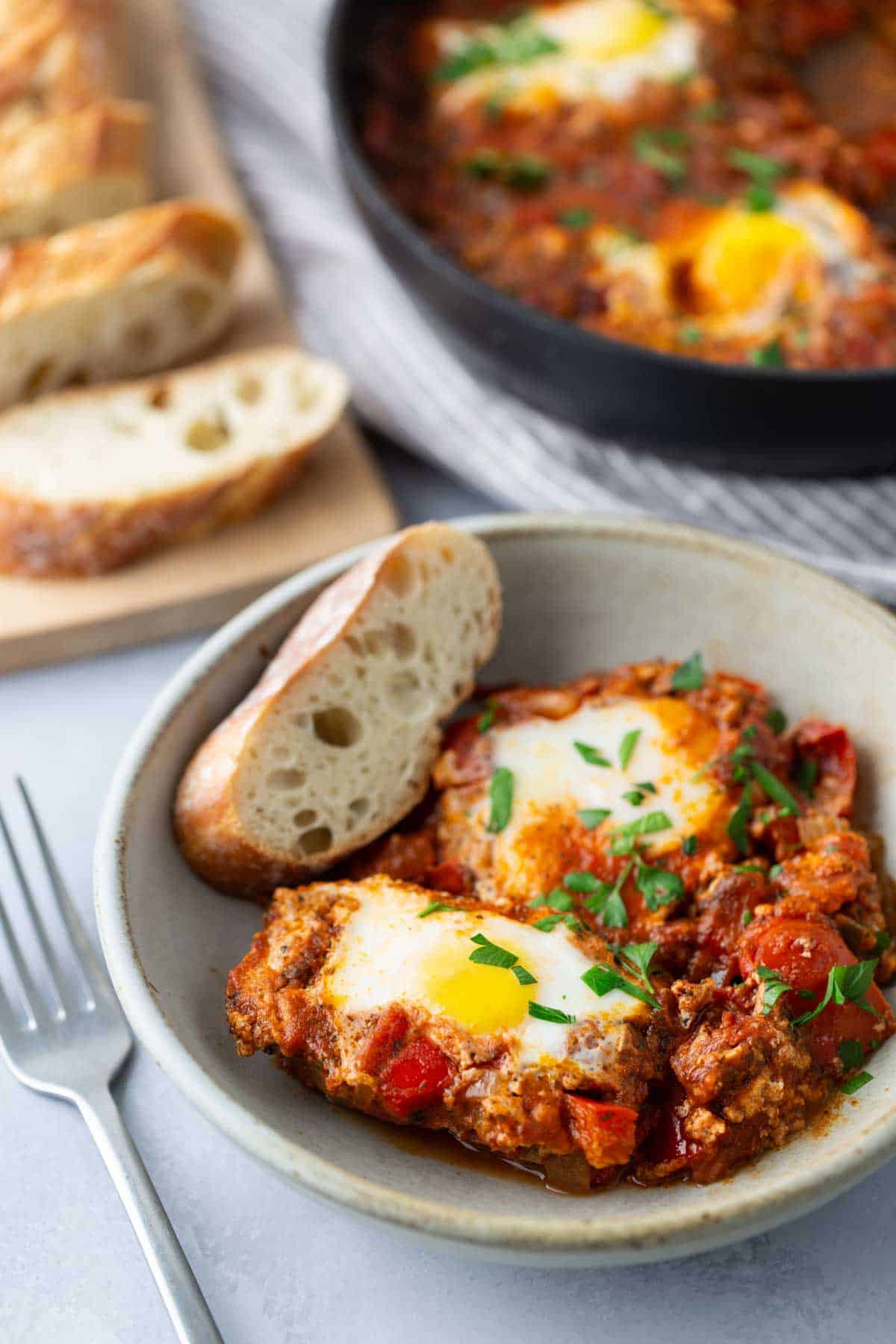 A bowl of shakshuka with two poached eggs in a tomato sauce, garnished with parsley, served with a slice of bread. A fork is placed beside the bowl, and more bread is on a cutting board in the background.