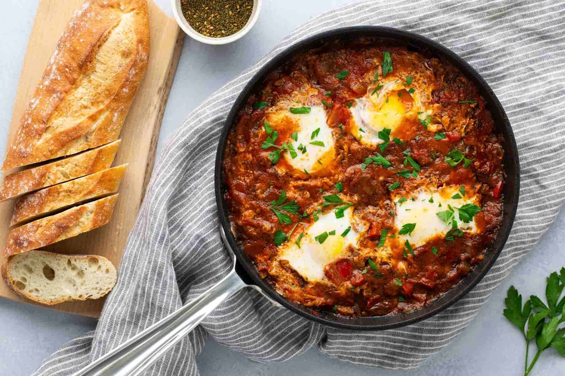 A skillet of baked eggs in a tomato-based sauce garnished with parsley, placed on a striped cloth next to a sliced loaf of bread on a wooden board.