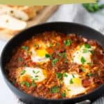 A skillet of za’atar shakshuka, featuring poached eggs in a tomato and pepper sauce, garnished with herbs. Slices of bread are visible in the background. Text overlay reads "Shakshuka.