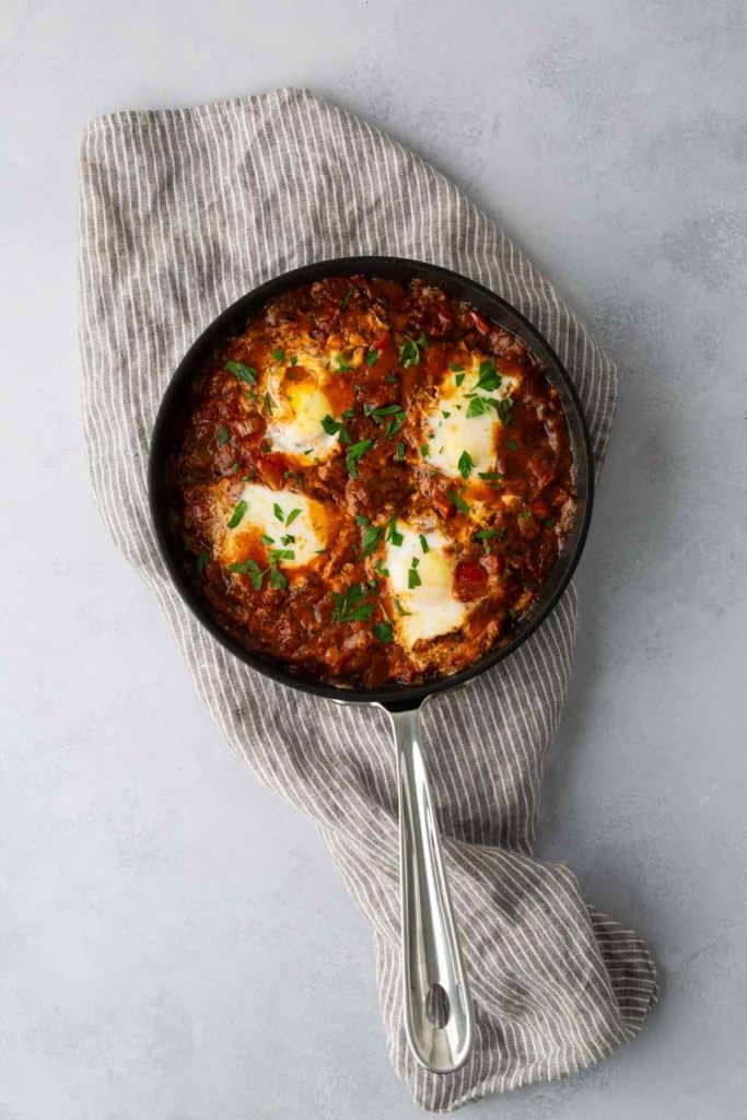 A black skillet filled with shakshuka, featuring poached eggs in a spiced tomato and pepper sauce, garnished with chopped parsley, resting on a striped cloth.