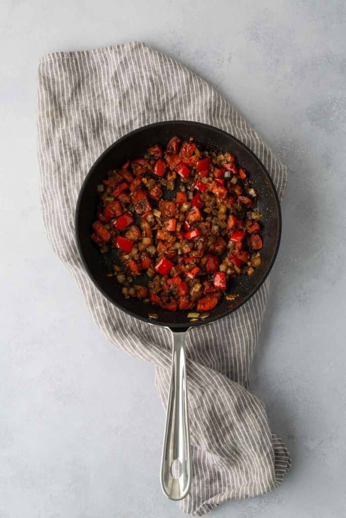 A black skillet with cooked diced vegetables, including red bell peppers and onions, rests on a gray and white striped cloth on a light-colored surface.