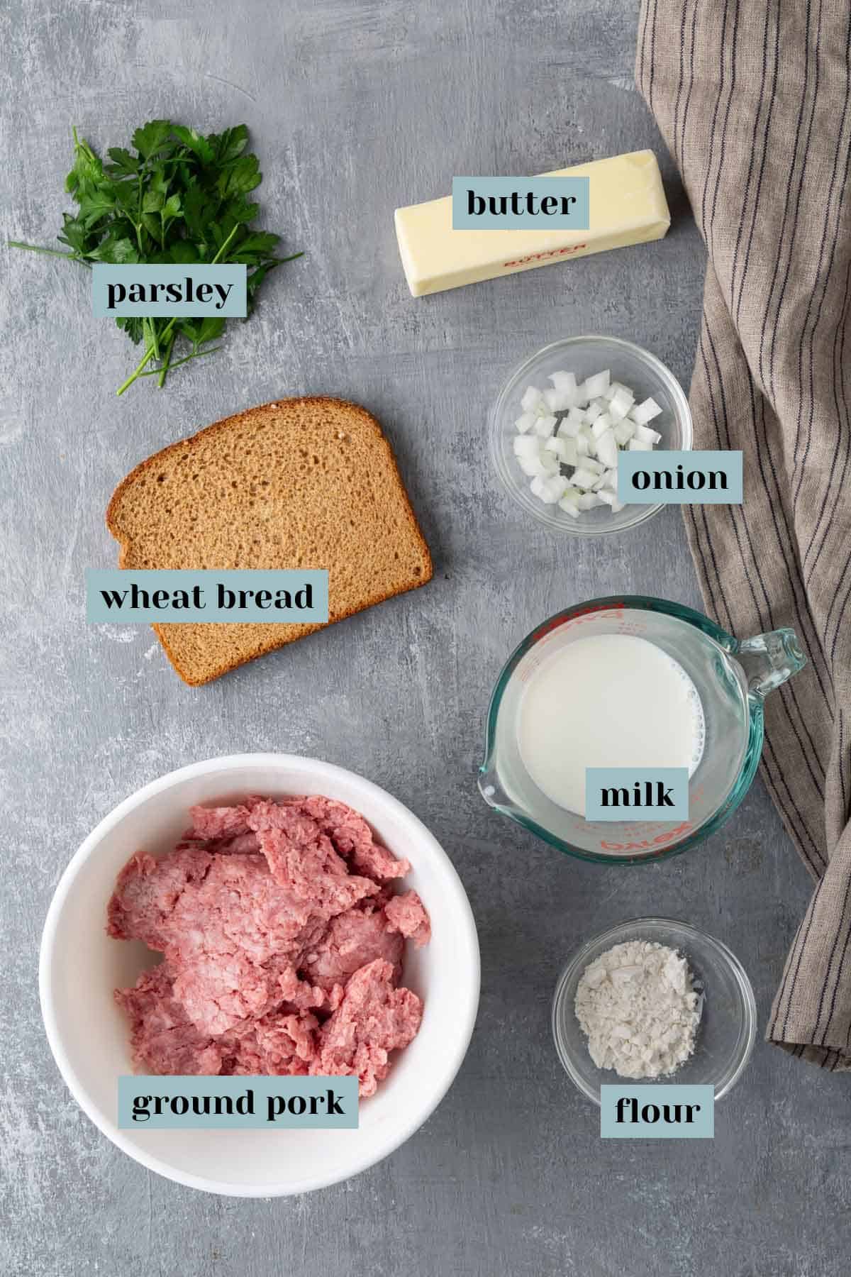 Ingredients including ground pork, milk, flour, onion, butter, parsley, and a slice of wheat bread laid out on a gray surface with a striped cloth beside them.