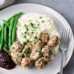 A plate with meatballs in sauce, mashed potatoes, green beans, and a dollop of red sauce, garnished with chopped herbs. A fork is placed on the side.