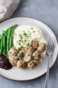 A plate with meatballs in sauce, mashed potatoes, green beans, and a dollop of red sauce, garnished with chopped herbs. A fork is placed on the side.