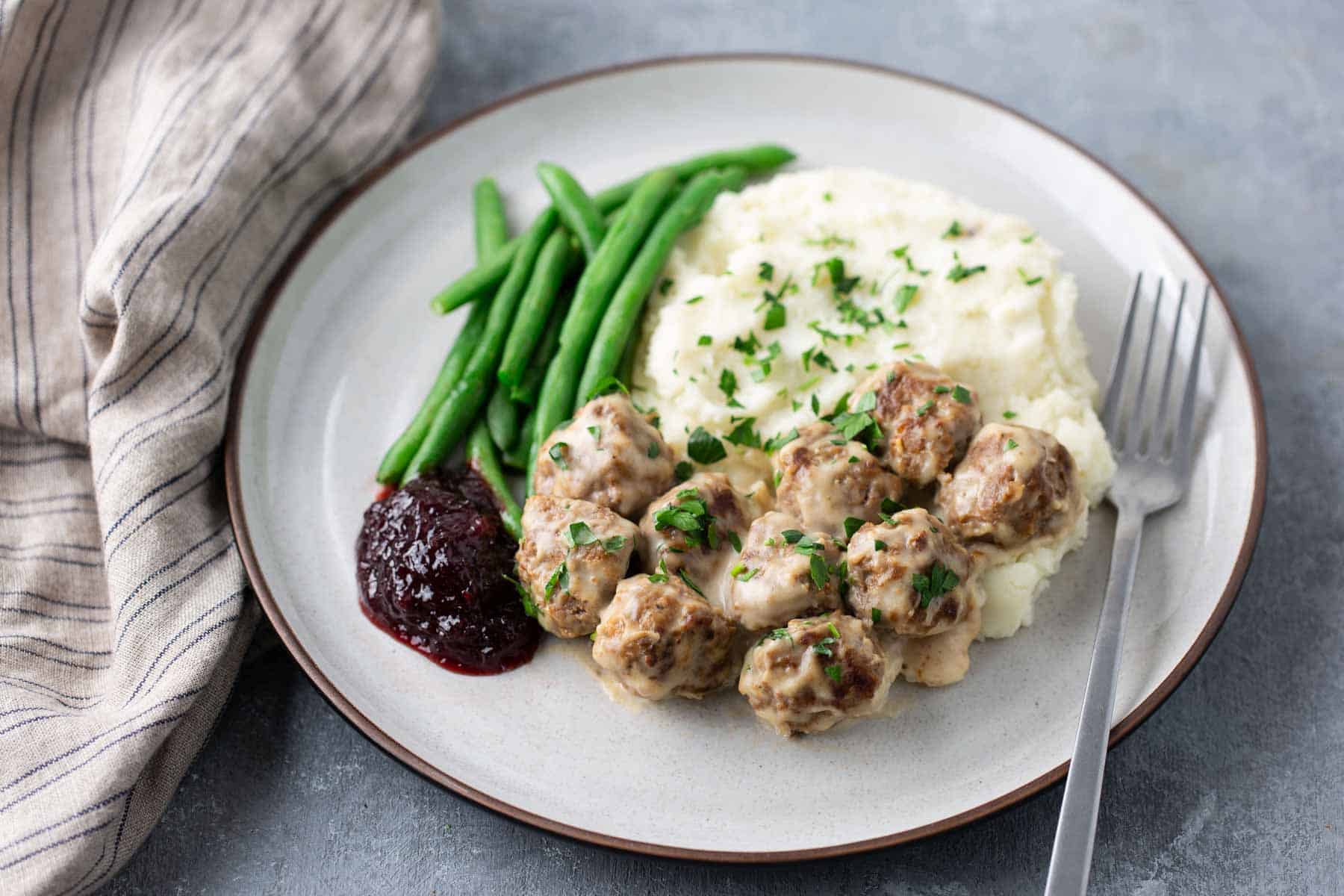 A plate of meatballs in gravy, green beans, mashed potatoes, and a dollop of lingonberry sauce, with a fork on the side. A striped cloth napkin lies next to the plate.