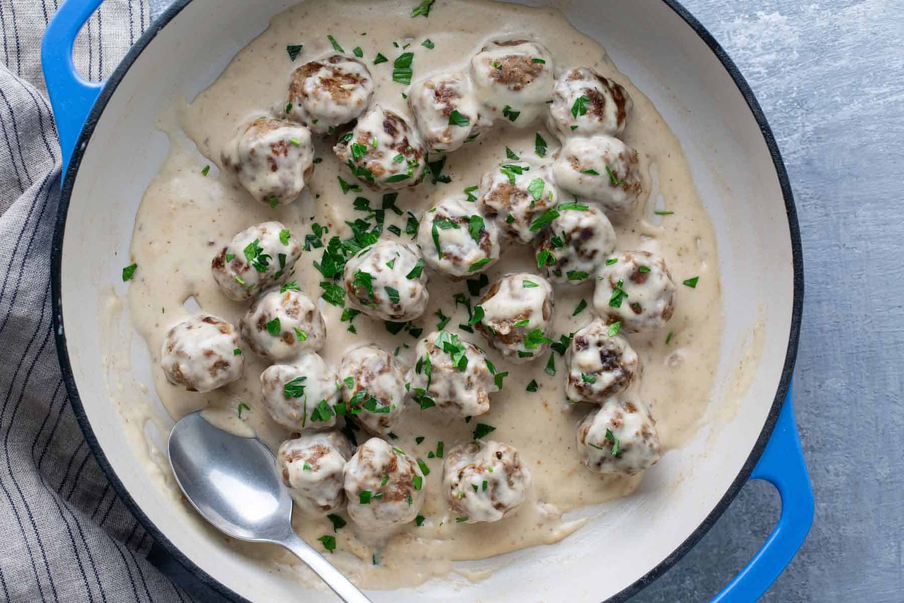 A pot of meatballs in creamy white sauce garnished with chopped parsley, with a silver spoon next to them.