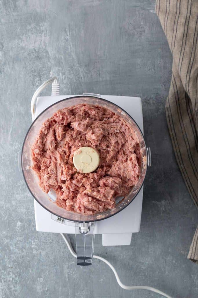 A food processor containing a mixture of ground meat sits on a grey countertop. A striped cloth is partially visible on the right side.