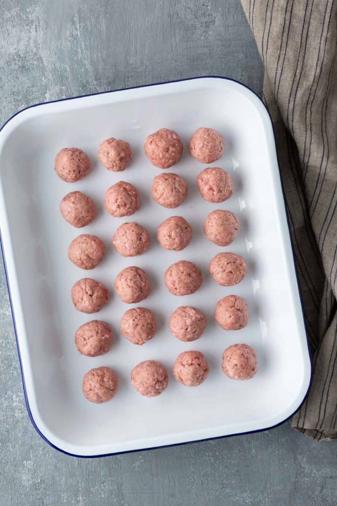 A white tray with 24 uniformly rolled raw meatballs on a gray surface, next to a folded striped cloth.