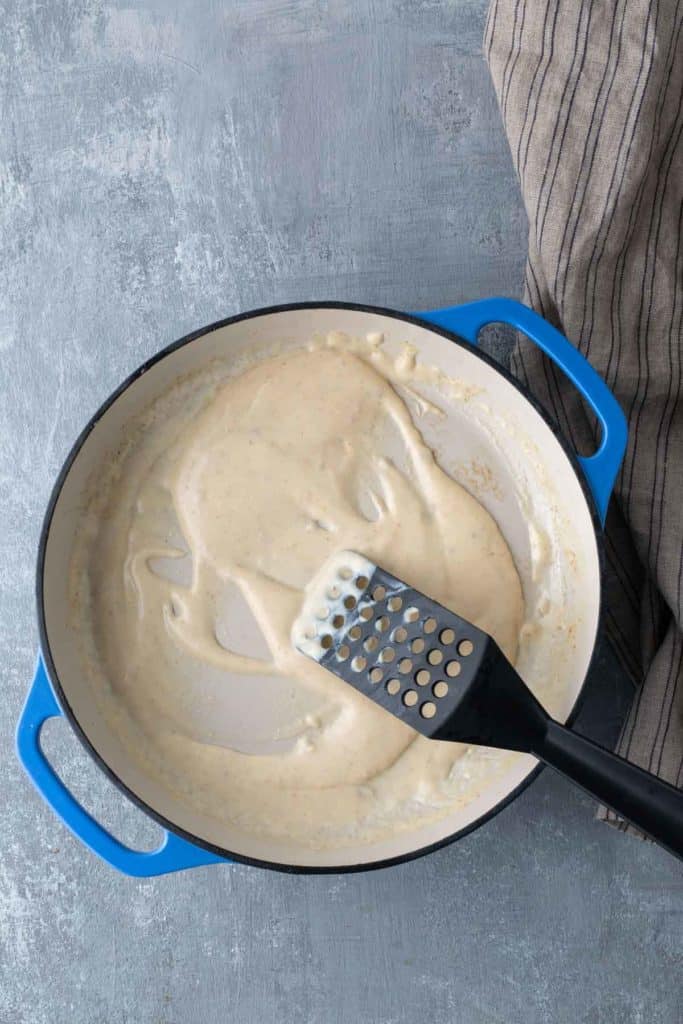 A blue-handled pot with a creamy white sauce being stirred by a black slotted spatula on a grey countertop next to a striped towel.