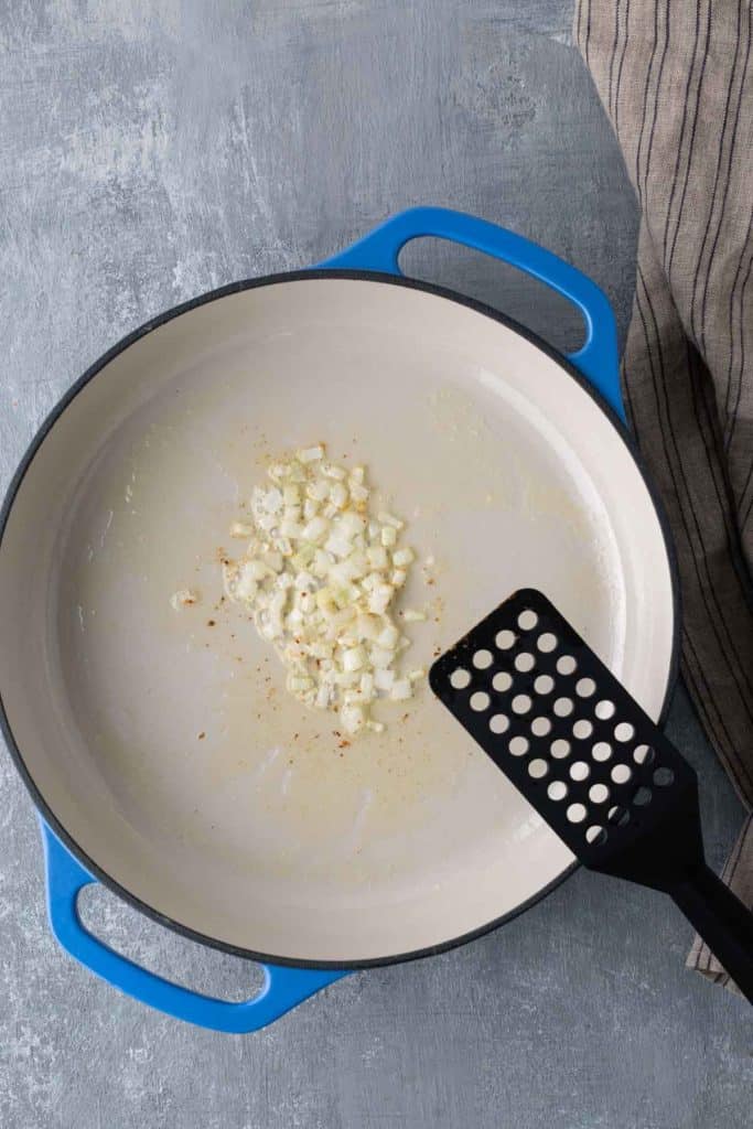 Chopped onions are sautéing in a large, blue-handled skillet with a spatula resting nearby on a grey surface.