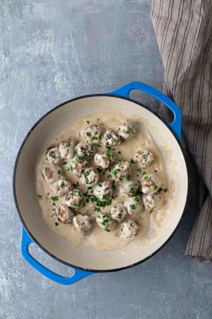 A blue-handled skillet containing meatballs in a creamy white sauce, garnished with chopped parsley, sits on a gray textured surface beside a striped cloth.