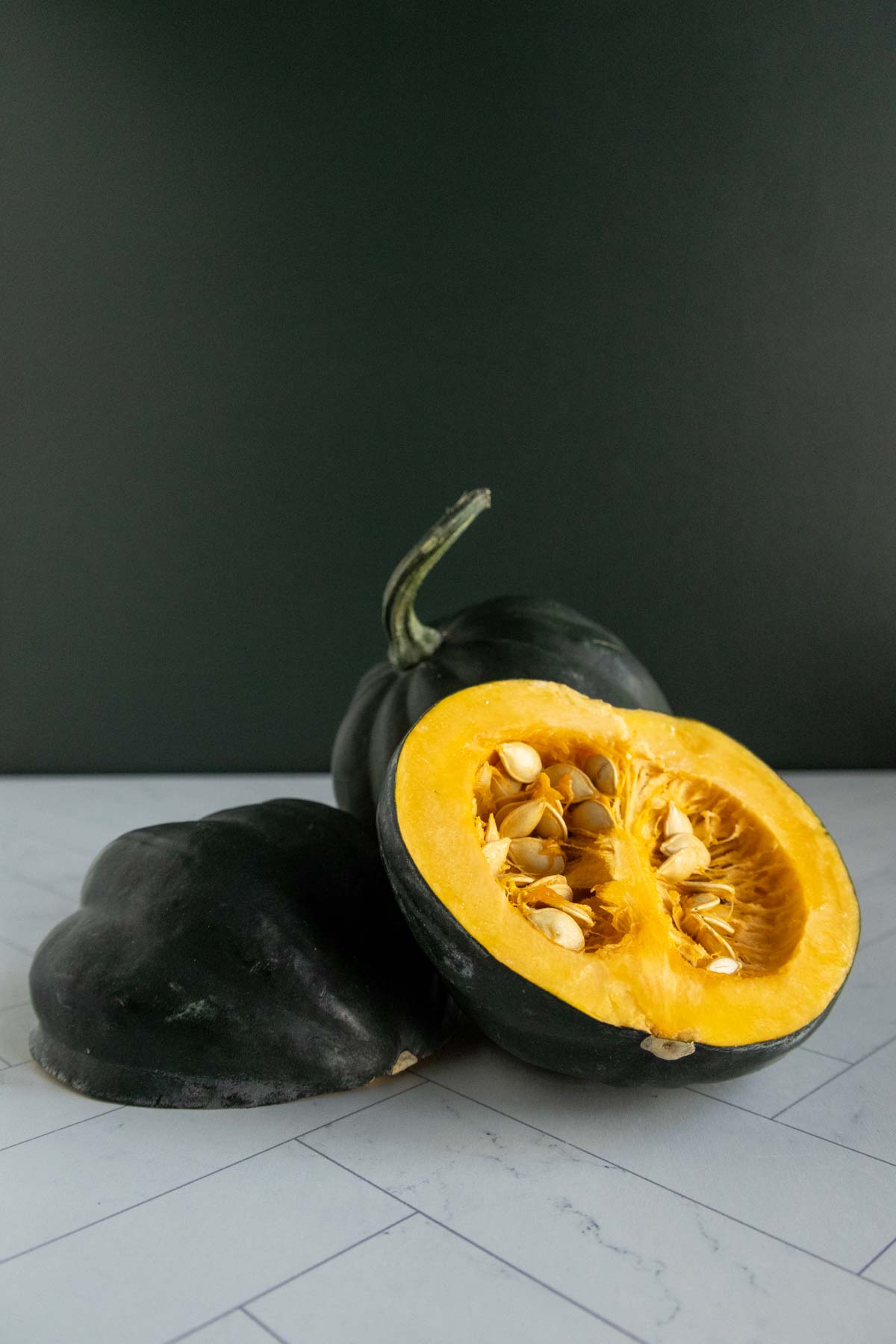 A dark green squash is cut open, revealing its bright orange flesh and seeds inside. Another piece of the squash lies beside it on a white surface with a dark background.