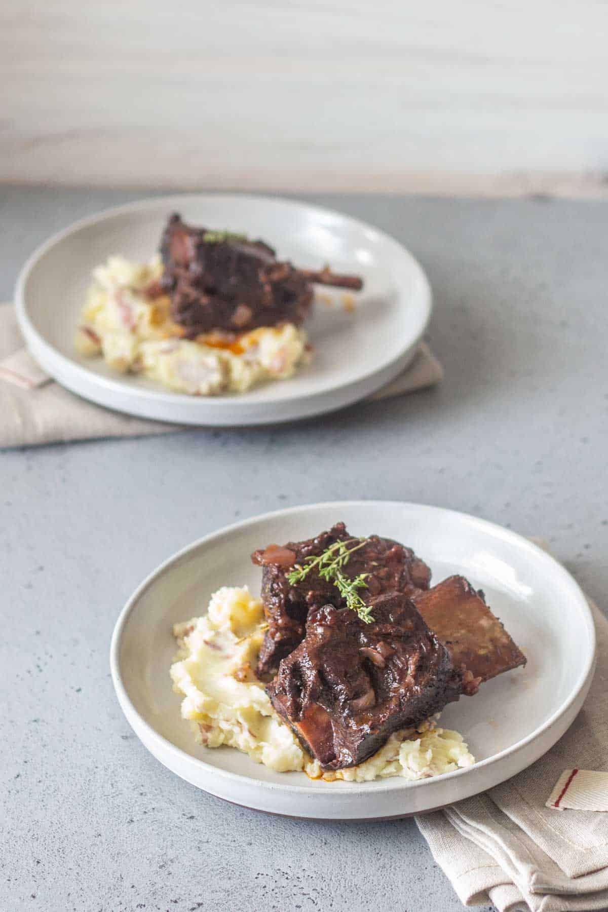 Two white plates, each with a serving of braised meat on a bed of mashed potatoes. The plate in the foreground showcases a sprig of thyme atop the meat. Both plates are placed on a gray surface.