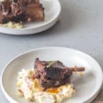 A plate with braised short ribs on top of mashed potatoes, garnished with herbs, and another similar plate in the background on a gray surface.