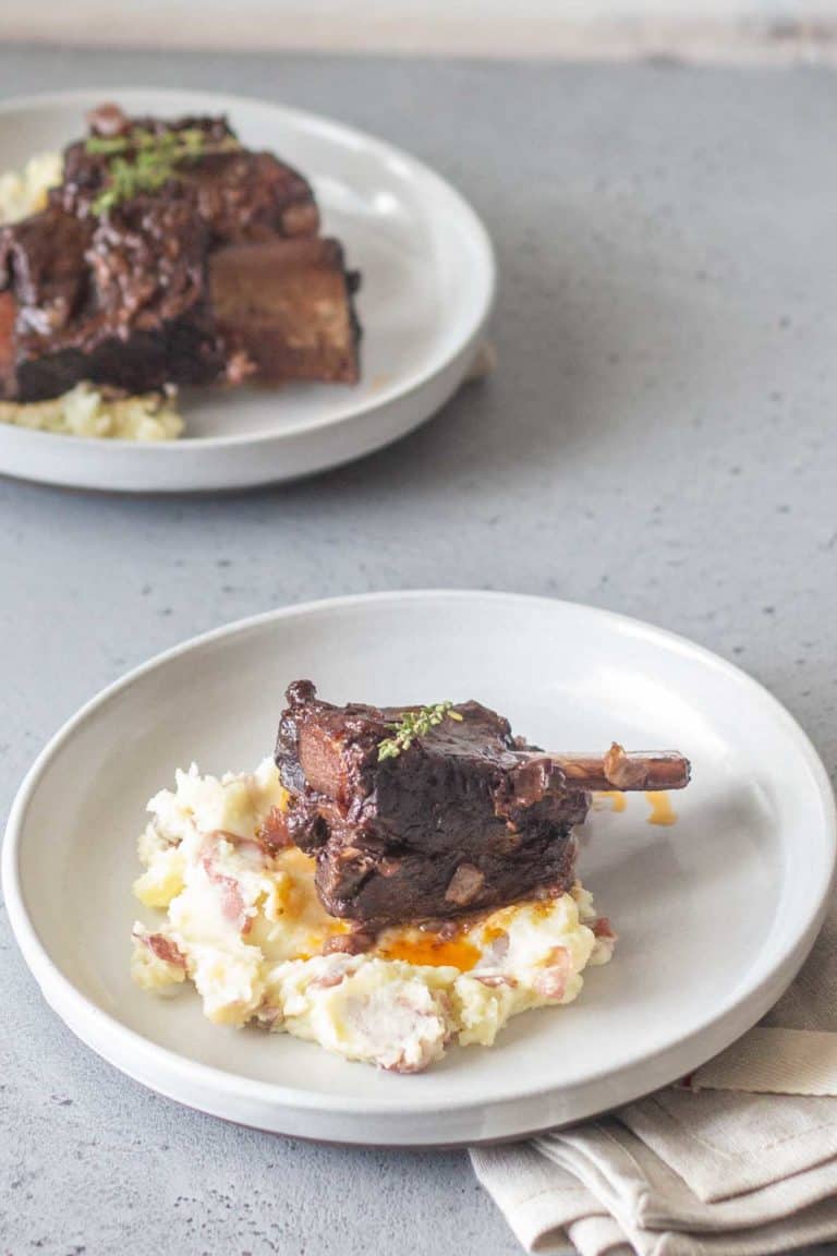 A plate with braised short ribs on top of mashed potatoes, garnished with herbs, and another similar plate in the background on a gray surface.