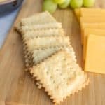 A wooden board with square crackers, slices of yellow cheese, and a bunch of green grapes.