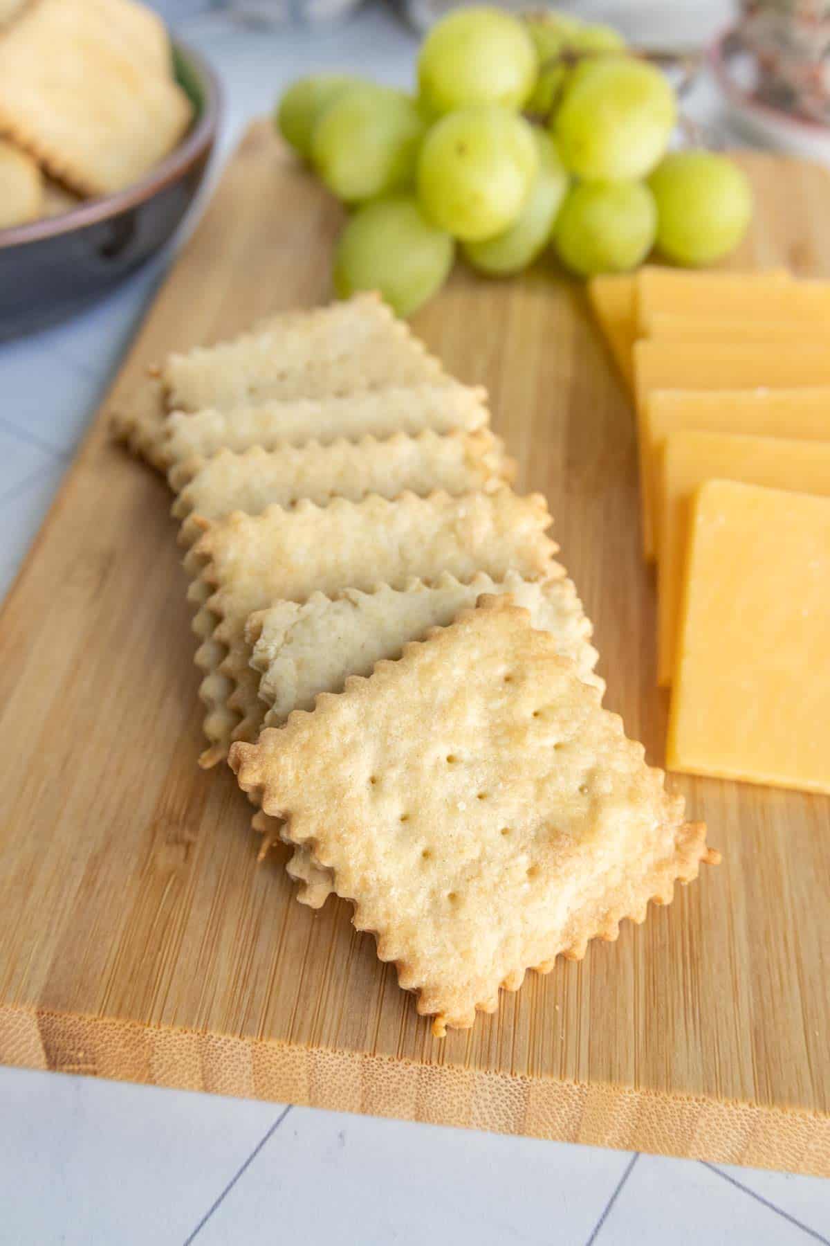 A wooden board with square crackers, slices of yellow cheese, and a bunch of green grapes.