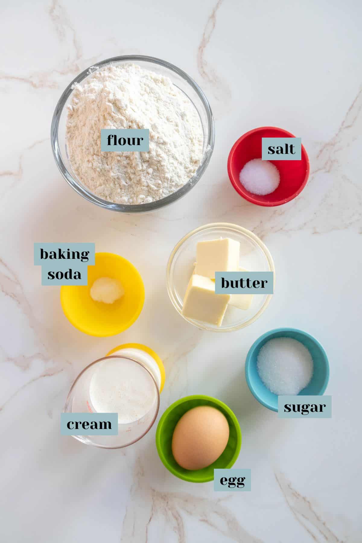 Overhead view of various baking ingredients in different colored bowls: flour, salt, baking soda, butter, sugar, cream, and an egg, arranged on a marble surface.