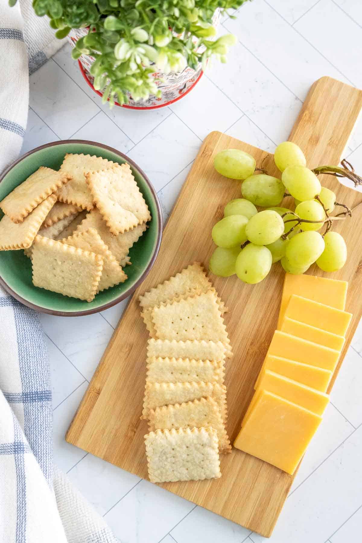 A wooden board with slices of cheddar cheese, green grapes, and rectangular crackers. A bowl of crackers and a small potted plant are placed nearby.