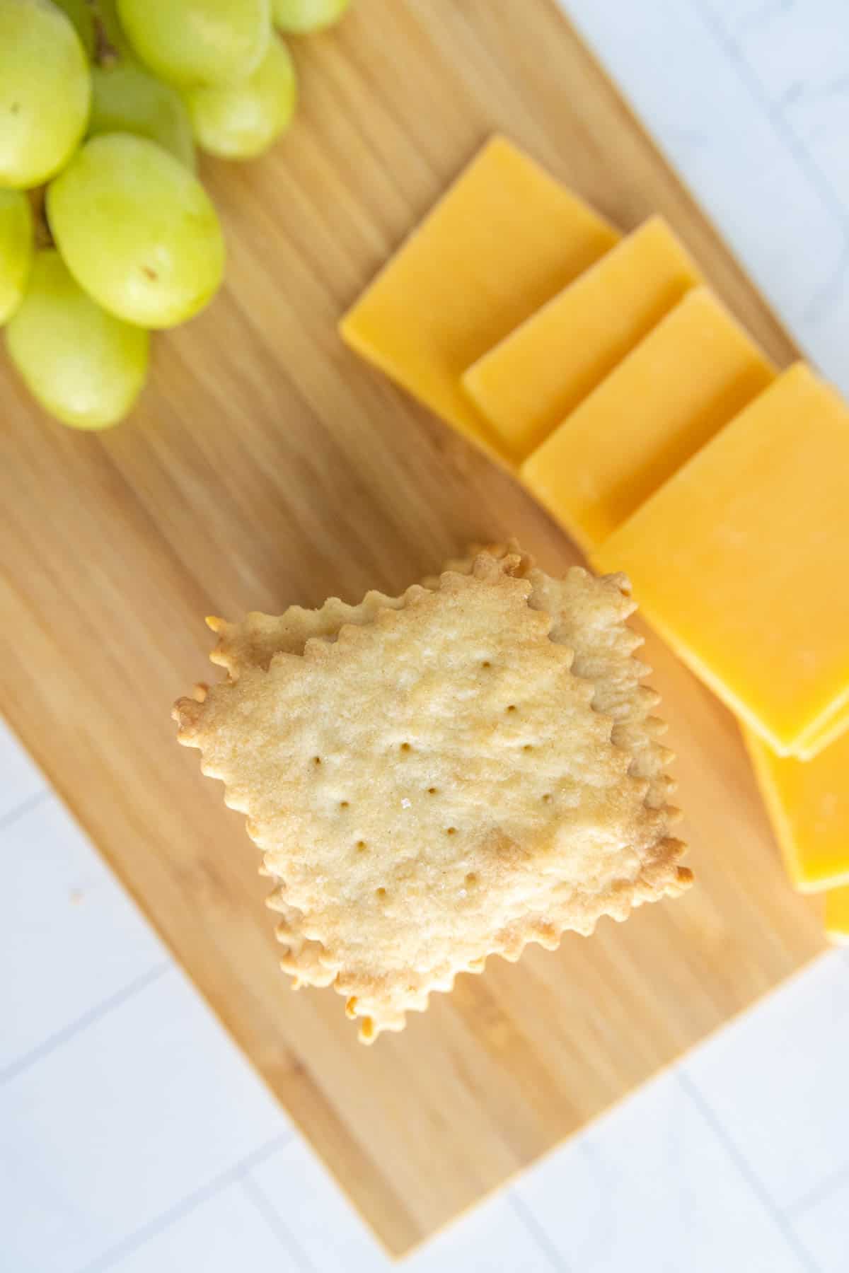 A wooden board with green grapes, sliced cheddar cheese, and square crackers.