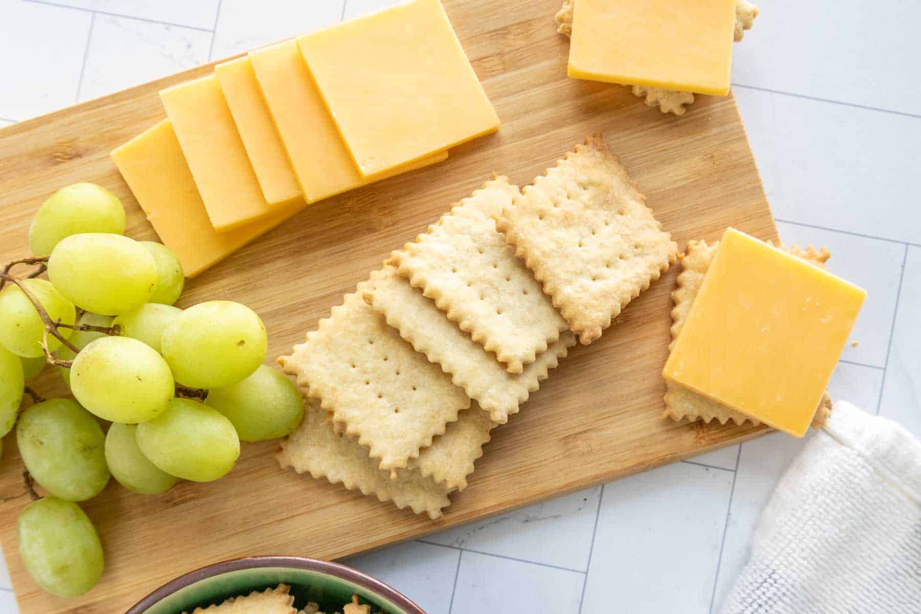 A wooden board with sliced cheddar cheese, crackers, and a bunch of green grapes. A kitchen towel is partially visible to the side.